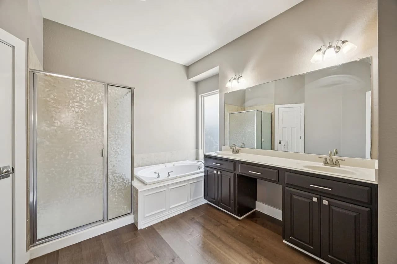 A rental bathroom as it is: gray walls, a glass shower, a white tub, a dark double vanity with brushed nickel faucets and a wide wall mirror.