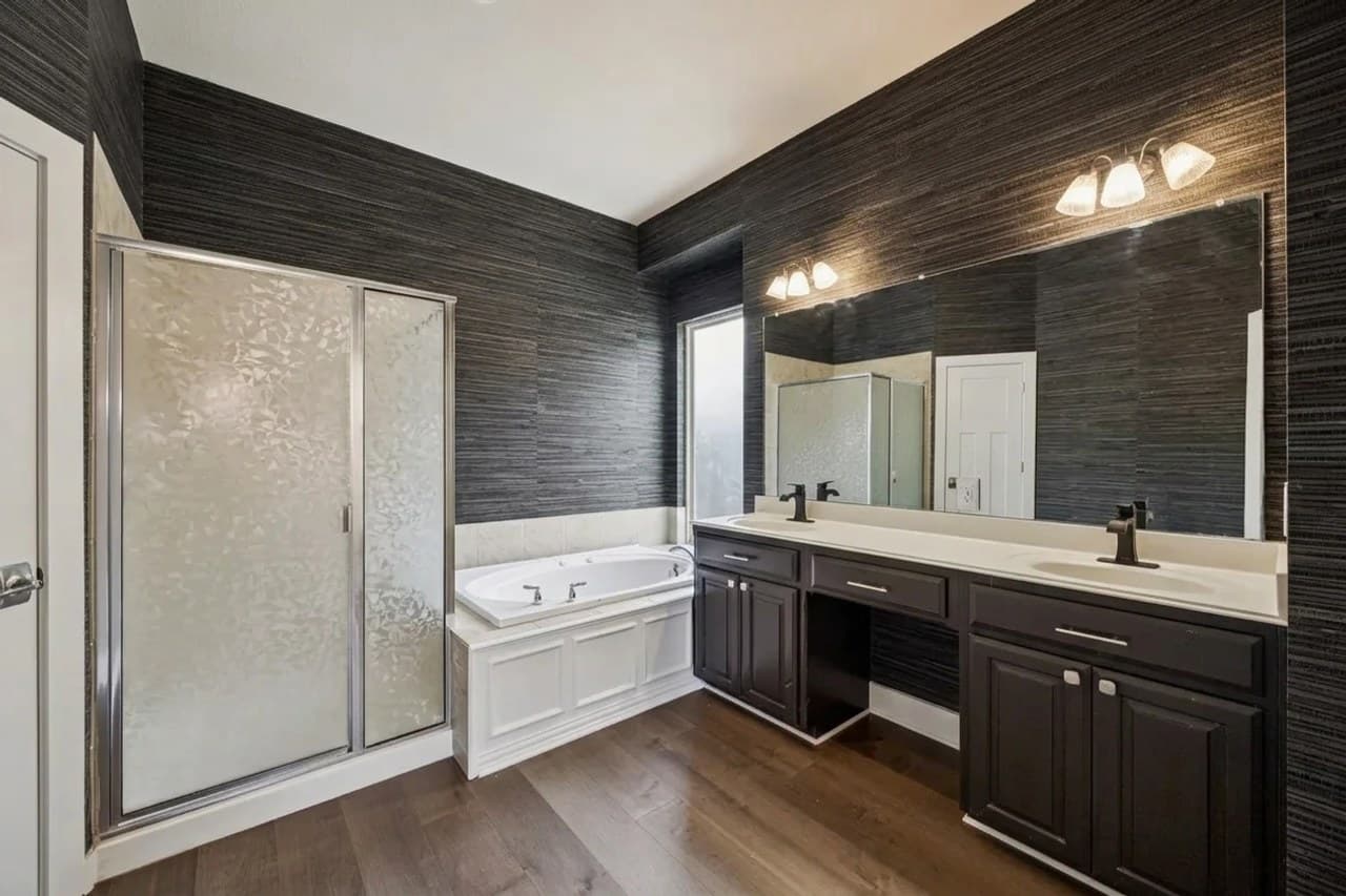 The same bathroom redesigned: charcoal grasscloth-look wallpaper on every wall and matte black faucets, with the shower, tub, vanity and floor unchanged.